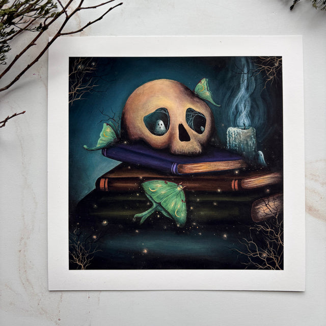 A square art print featuring a surreal illustration of a skull resting on stacked books with green butterflies and a smoky candle, displayed on a light marble surface with decorative branches in the corners.