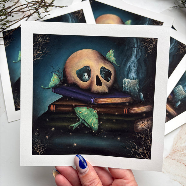A hand with blue and gold nail polish holds a square art print featuring a surreal illustration of a skull resting on stacked books with green butterflies in a dark blue scene. Other similar prints are fanned out behind it on a light marble surface with dried botanicals in the corner.