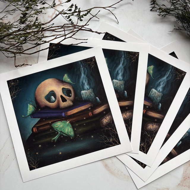 A set of illustrated art prints with white borders, fanned out on a light marble surface with dried botanical stems. The artwork depicts a skull resting on books with green leaves and a dark smoky background.