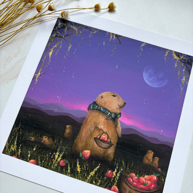 A square art print featuring a whimsical illustration of a prairie wearing a green plaid scarf and holding a basket of peaches in a field at night under a purple and pink moonlit sky. The print is displayed on a light surface with dried flowers in the corner.