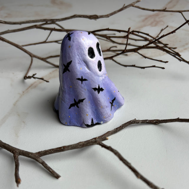 A small ghost-shaped figurine painted in light lavender purple with black bat and eye details, standing on a white surface with bare branches around it.