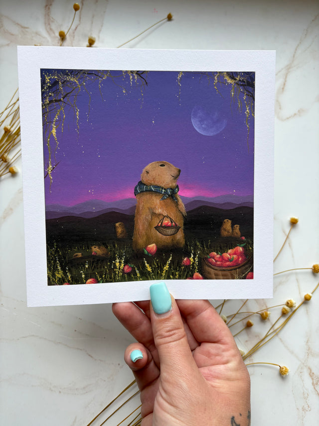 A hand with light blue nail polish holds a square art print featuring a whimsical illustration of a praires dogs wearing a green scarf and holding baskets of apples under a purple night sky with a crescent moon. The print is displayed on a light marble surface decorated with dried flowers.