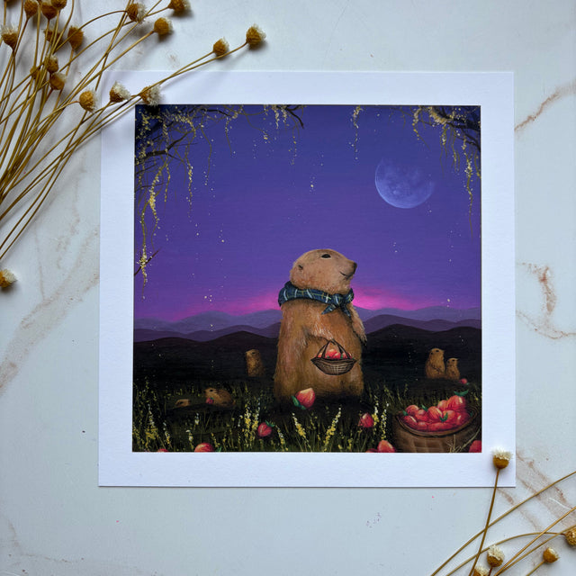 A square art print featuring an illustrated prairie dog standing in a field at night under a purple sky with a moon, displayed on a light marble surface with dried flowers around it.