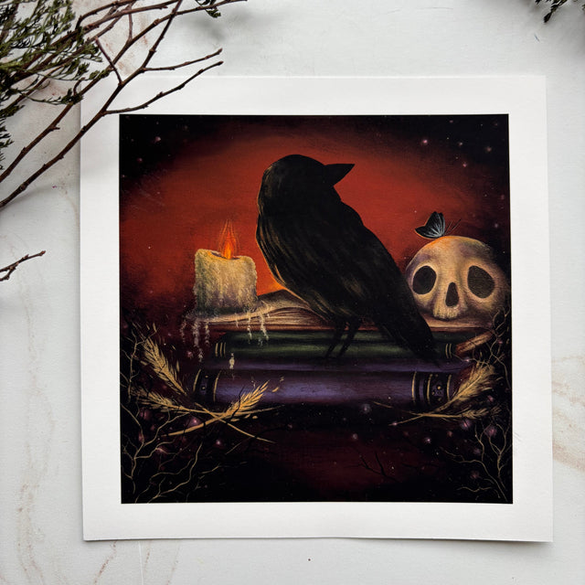 A square art print with a white border featuring a dark gothic illustration of a black raven perched on stacked books beside a skull and glowing candle, set against a red and black background. The print is displayed on a light marble surface with dried botanical branches in the corners.