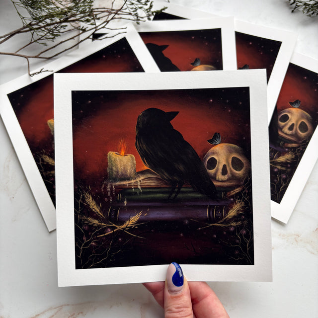 A hand with blue nail polish holds a square art print featuring a black raven perched on a book beside a skull and candle against a dark red and black background. Several matching prints are fanned out behind it on a light marble surface with dried greenery in the corner.